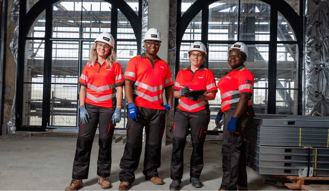 Four construction workers in red and black uniforms, white hard hats, and protective gloves stand and pose inside a building under construction.