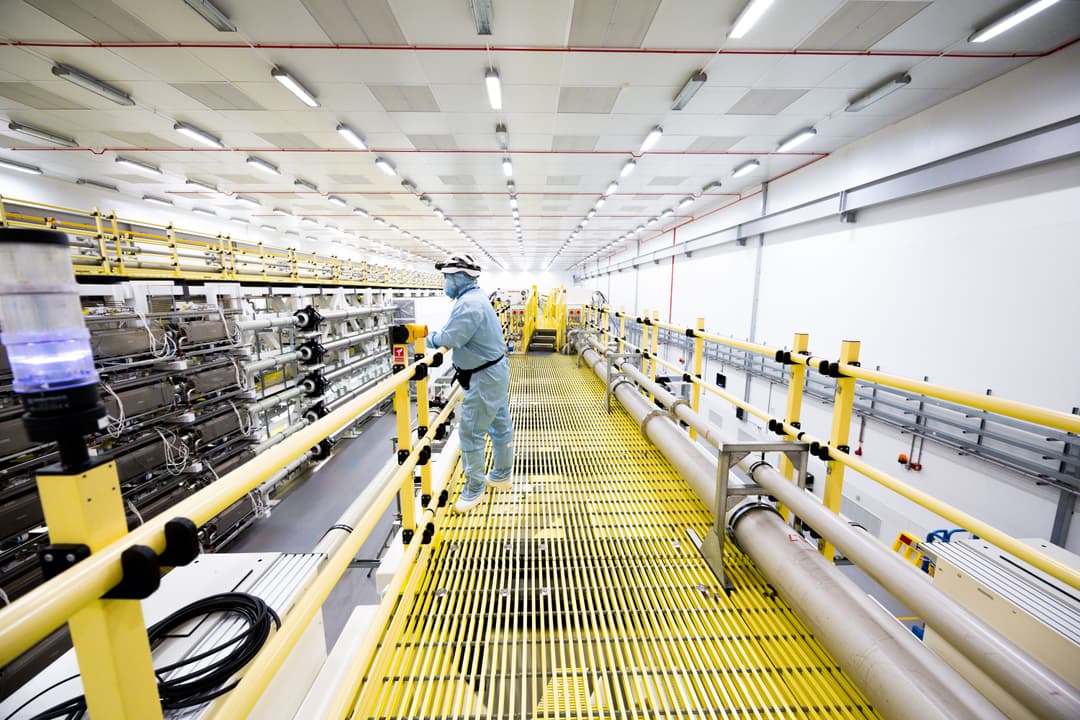 Worker in full protective clothing standing on a yellow grated walkway inside a large, brightly lit industrial facility with pipes and equipment on both sides
