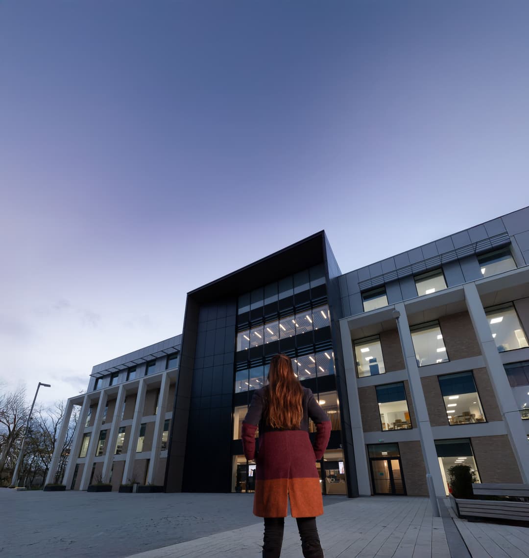 Person in a long coat standing with their back to the camera, facing a modern office building at dusk