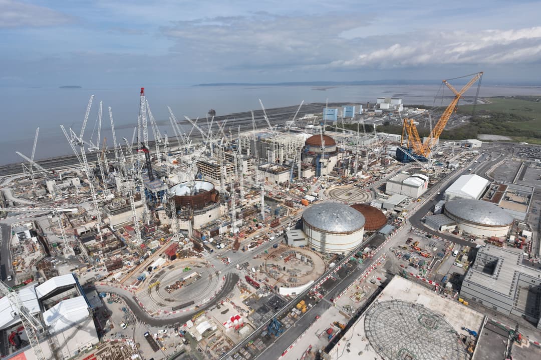 Aerial view of a large nuclear power station construction site with multiple domed reactor buildings, cranes and surrounding infrastructure under a cloudy sky