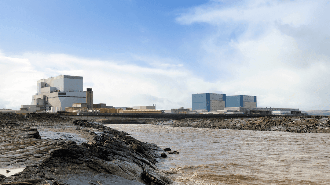 Low shoreline with rocky outcrops in the foreground and large industrial buildings of a power station across the water under a partly cloudy sky