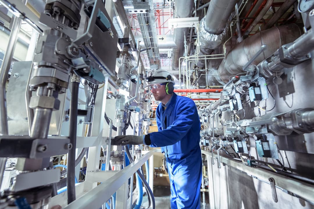 Technician in blue coveralls and protective gear inspecting industrial piping and valves inside a complex energy facility