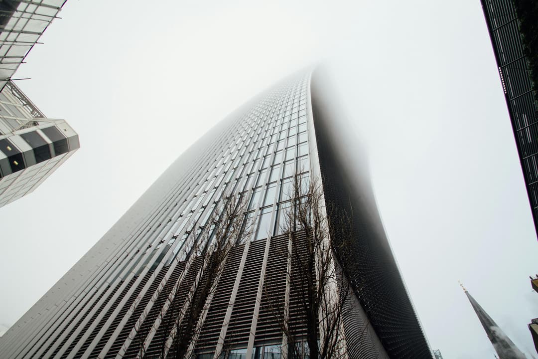 Low-angle view of a modern glass and steel office tower with a curved façade rising into a cloudy sky, flanked by other buildings and bare tree branches at the base.